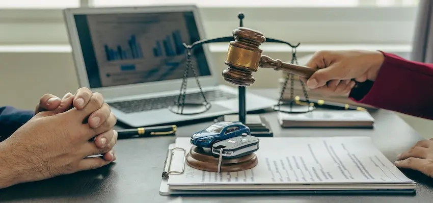 Two people sit across from each other at a dark wooden desk engaged in a formal legal consultation with the person on the left resting both hands clasped together on the table surface while the person on the right in a red sleeve reaches across to hold a wooden judge's gavel with a gold band over a round sound block beside which a small blue toy car sits on top of a set of metal car keys- a clipboard holding a signed legal document in the foreground- a brass balance scale of justice standing upright between them- and an open laptop displaying a blue bar graph visible in the background — illustrating the evidence review and settlement valuation meeting that takes place when determining what a car accident case is worth at South Carolina Car Accident Lawyers.