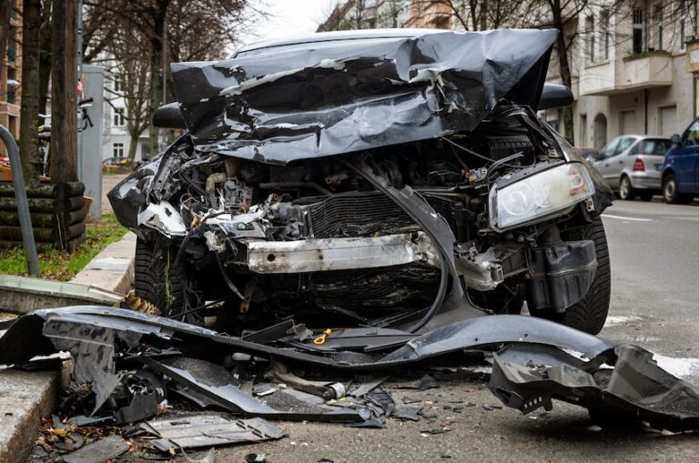 A black sedan has been destroyed from the front end in a high-impact collision- photographed head-on from the street level on a gray overcast day with the entire hood folded and crushed downward and backward toward the windshield exposing the full engine compartment, the radiator- front axle components- and internal mechanical parts all visible and severely deformed large jagged pieces of black plastic bumper and body panels scattered across the concrete curb and sidewalk in the foreground- one surviving headlight unit still intact on the right side of the frame and a European urban streetscape with parked vehicles- residential buildings- and bare winter trees lining the road visible in the background — the kind of total-loss vehicle destruction that demands immediate legal representation to build a strong car accident case in South Carolina.