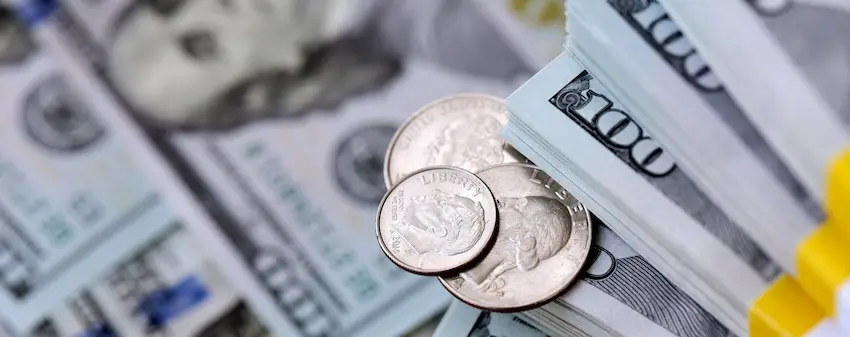 A tightly cropped close-up image shows two silver American quarters with "LIBERTY" and the eagle emblem visible on their faces resting on top of a thick neatly banded stack of crisp hundred-dollar bills secured with a yellow band- the bold black numeral 100 printed on the edge of the stack clearly legible with additional loose hundred-dollar bills fanned out beneath in a soft blur filling the entire background of the frame — a detailed representation of the financial compensation that injury victims seek to recover for damages- medical expenses- and lost income when pursuing a car accident case with help from South Carolina Car Accident Lawyers in South Carolina.